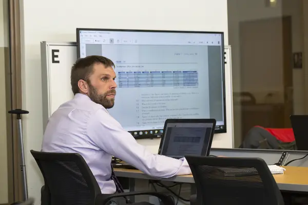 Male IT student at a desk with a laptop, screen displaying spreadsheet data.
