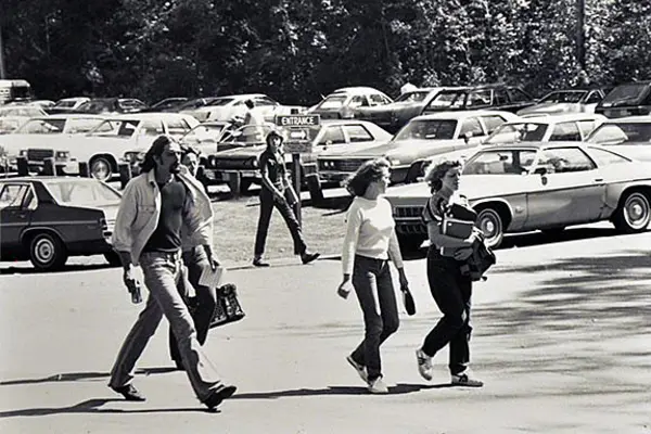 Students walking in campus parking lot with vintage cars in the background, black and white photo.