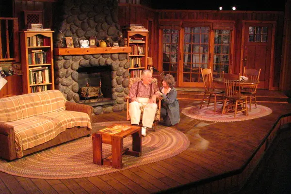 The Nicolet Players performing On Golden Pond. Cozy living room set with wood decor, a fireplace, and two people sitting.