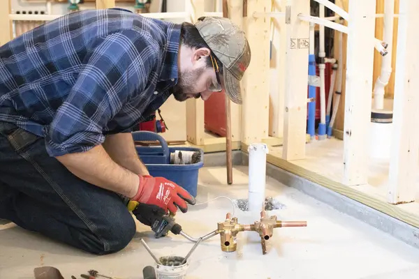 Male plumber working on copper pipes in a construction site, wearing a plaid shirt and cap.