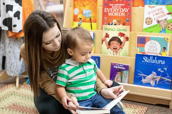 Early Childhood Education student and toddler reading a book in a colorful library setting.