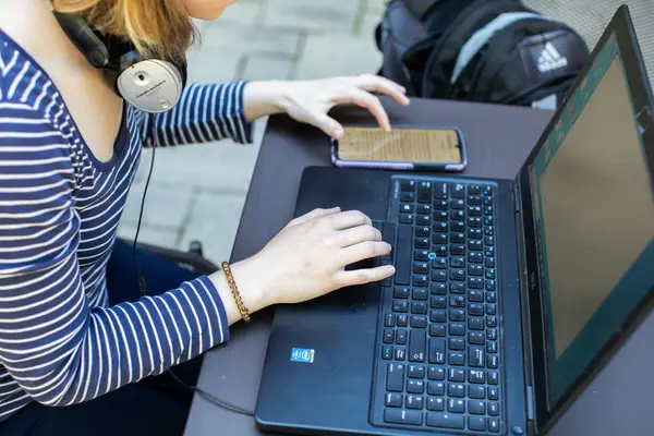 Person using a laptop and smartphone outdoors with headphones.