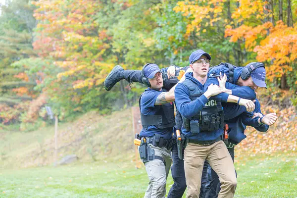 Recruits training exercise with three men carrying another in a grassy, autumn setting.