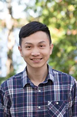 Young man smiling in a plaid shirt, with a blurred green background.