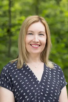 Woman smiling in a forest setting, wearing a navy patterned blouse.