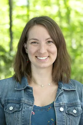 Smiling woman in denim jacket, outdoors with green foliage background.