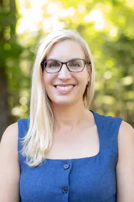 Smiling woman with glasses, blonde hair, wearing a blue dress, outdoors.