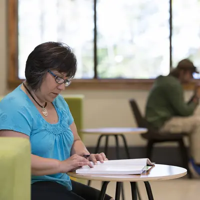 Female student reading at a table in a bright, calm room; another person in background on a laptop.