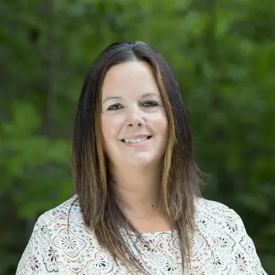 Smiling woman in patterned top, green leafy background.