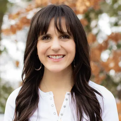 Smiling woman with long hair, white shirt, autumn leaves in background.