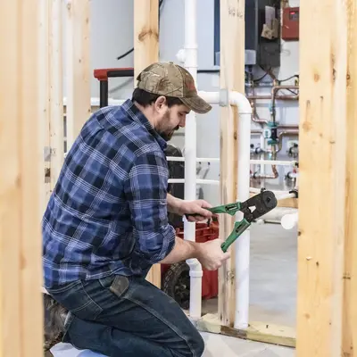 Plumbing apprentice kneeling, using green pliers on pipes in a construction area.