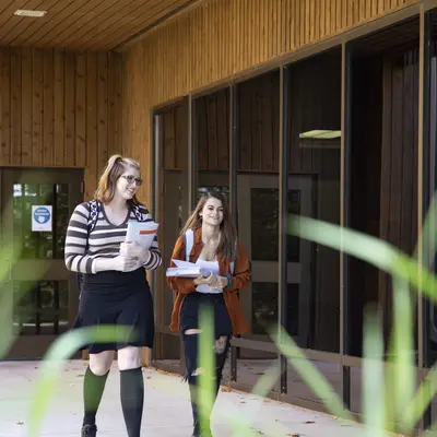 Two female students walking outside a wooden building, holding papers, smiling.