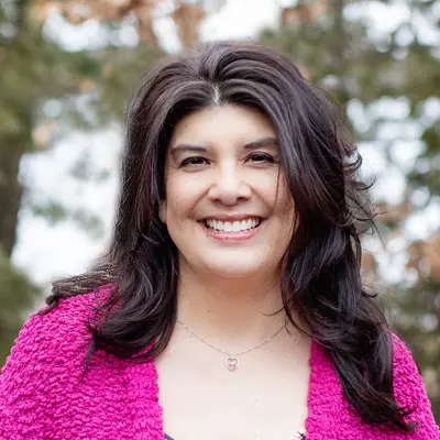Smiling woman in a pink cardigan outdoors with blurred trees.