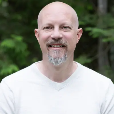Man with a goatee smiling in a white shirt, green foliage background.