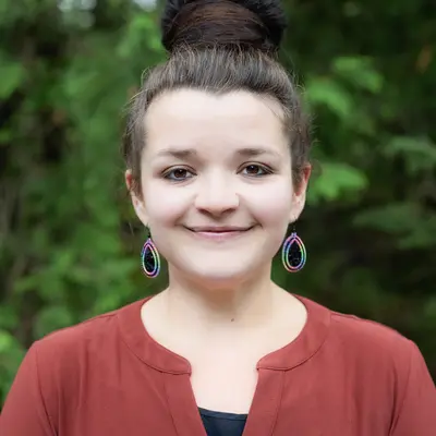 Young woman smiling, wearing a red blouse, with greenery in the background.