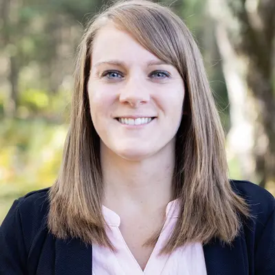 Smiling woman outdoors, wearing a blazer and pink shirt.