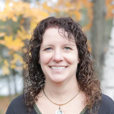 Smiling woman outdoors, wearing a necklace, with autumn leaves in the background.