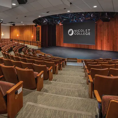 Theatre with rows of brown seats facing a stage and projector screen reading "Nicolet College."