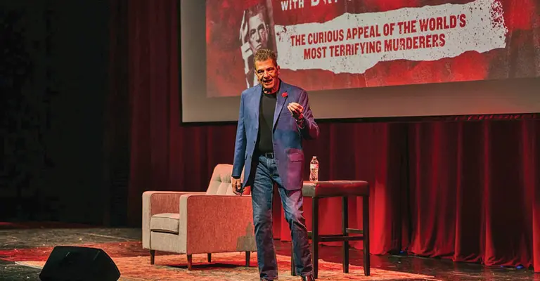 Speaker on stage with a table, chair, and red curtains in the background.