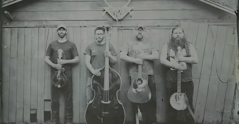 Four musicians with string instruments stand in front of a wooden fence, black and white photo.