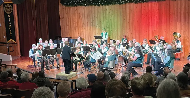 Concert band performing on stage with audience seated, festive lights above.