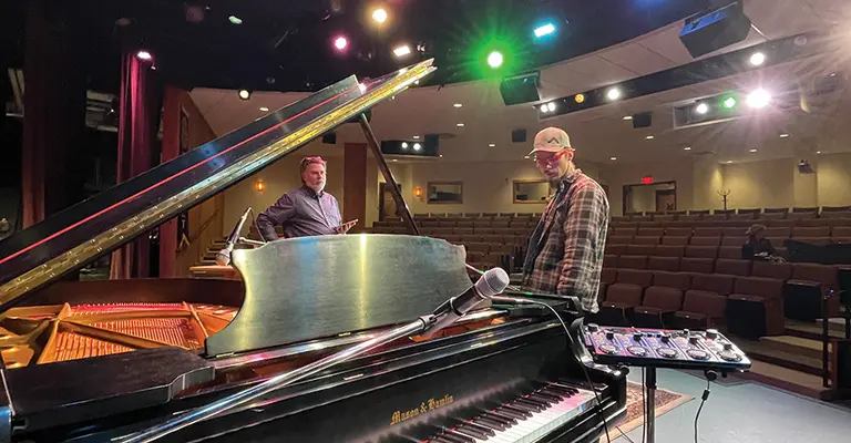 Grand piano on stage under colorful lights with two people in the background.