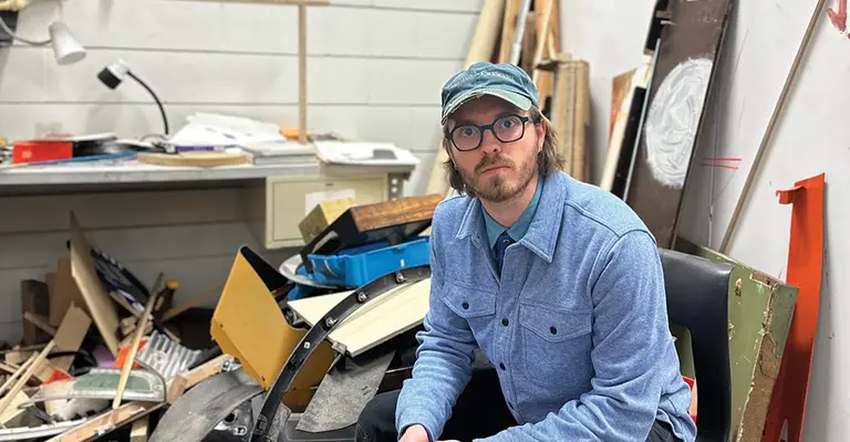 Artist in a workshop sits near scattered tools and wood materials.