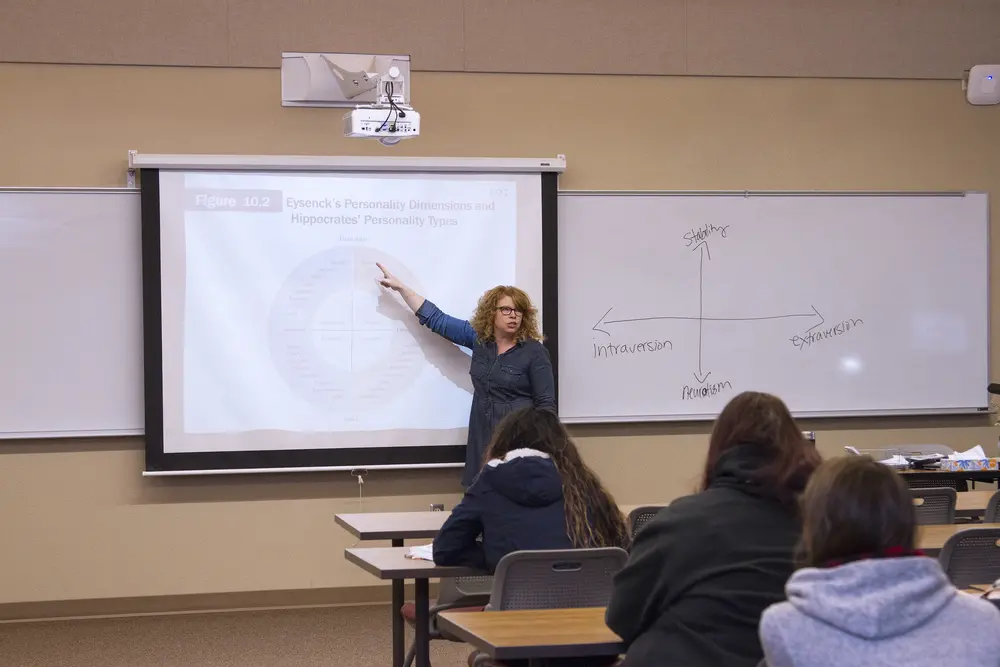 Female instructor pointing at a projector screen in a classroom with students.