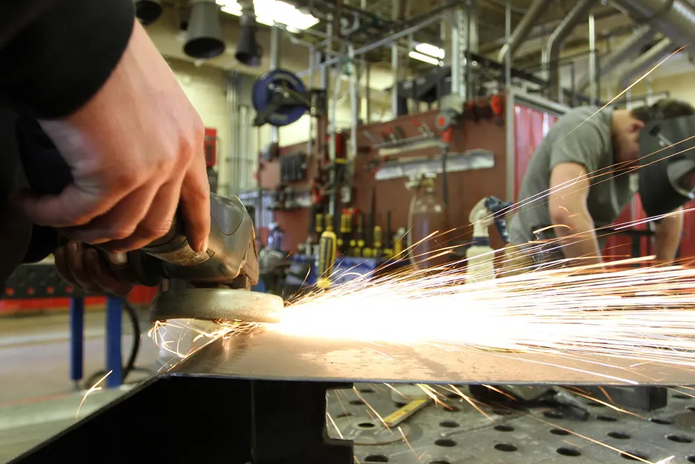 Student using a grinder, sparks flying, in an industrial workshop.