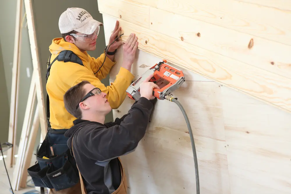 Two male carpentry apprentice students working on wood paneling with a nail gun.