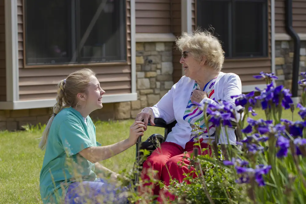 Female CNA student and elderly female patient in wheelchair smiling in garden at a healthcare facility.