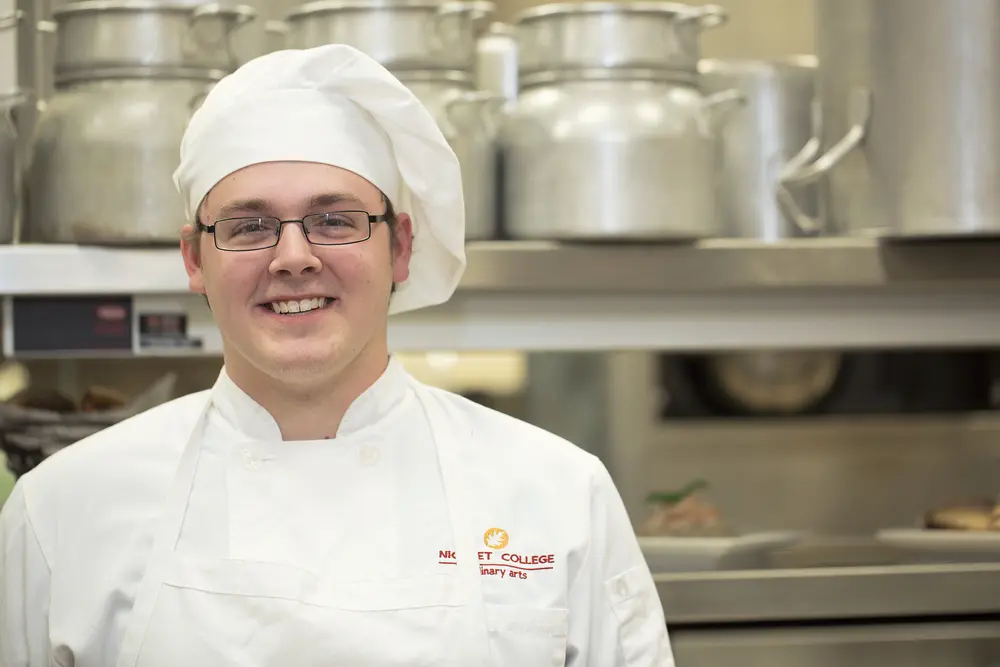 Male culinary student smiling in a kitchen wearing a white uniform and hat.