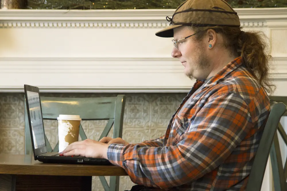 Male online student in a plaid shirt typing on a laptop in a cafe, coffee cup nearby.