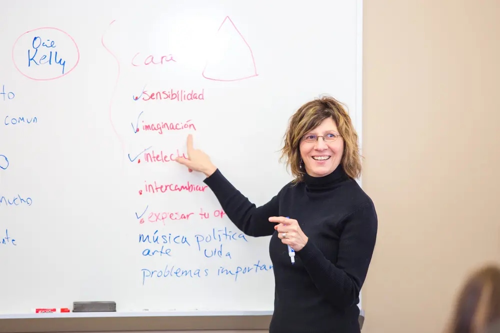 Spanish instructor smiling and gesturing at a whiteboard with colorful writing.