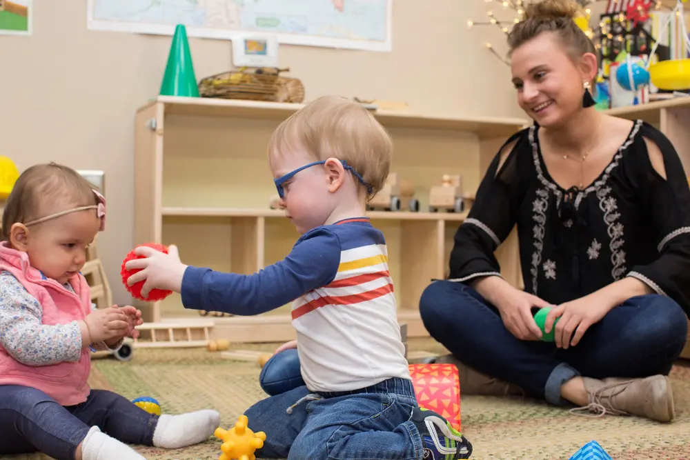 Two toddlers play with a ball, supervised by a student Early Childhood Education teacher.