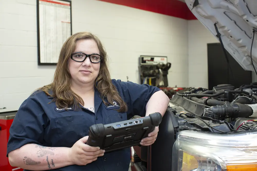 Female Automotive student holding a diagnostic tool by an open car hood.