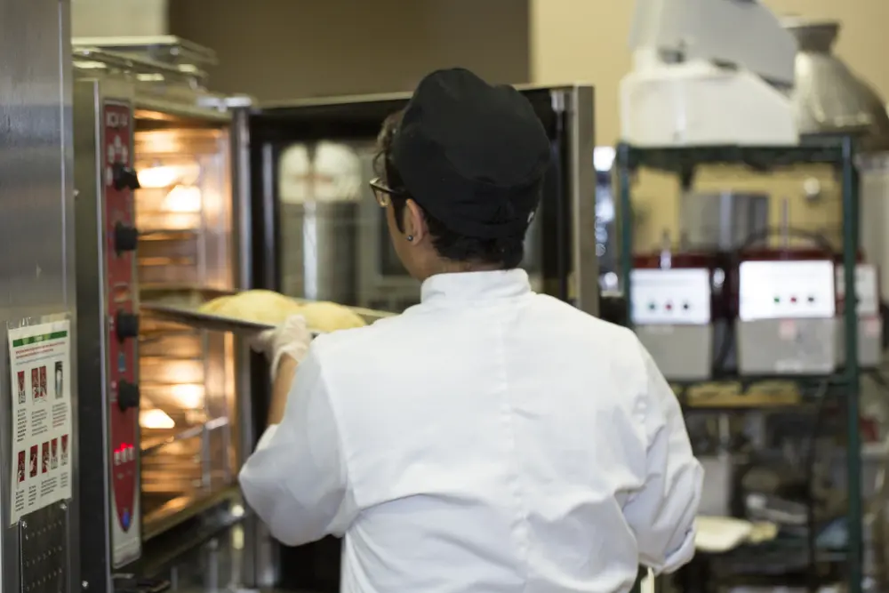 Culinary student placing tray of bread in an oven in a commercial kitchen.