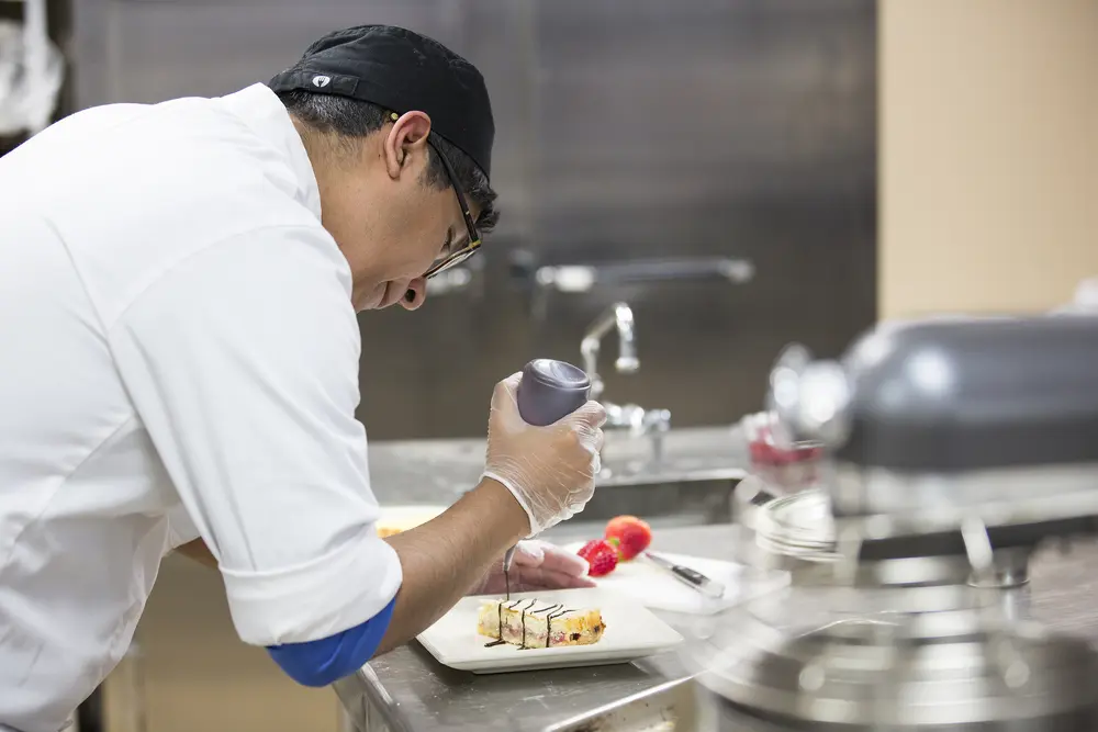 Culinary student focused on plating a dessert in a kitchen.