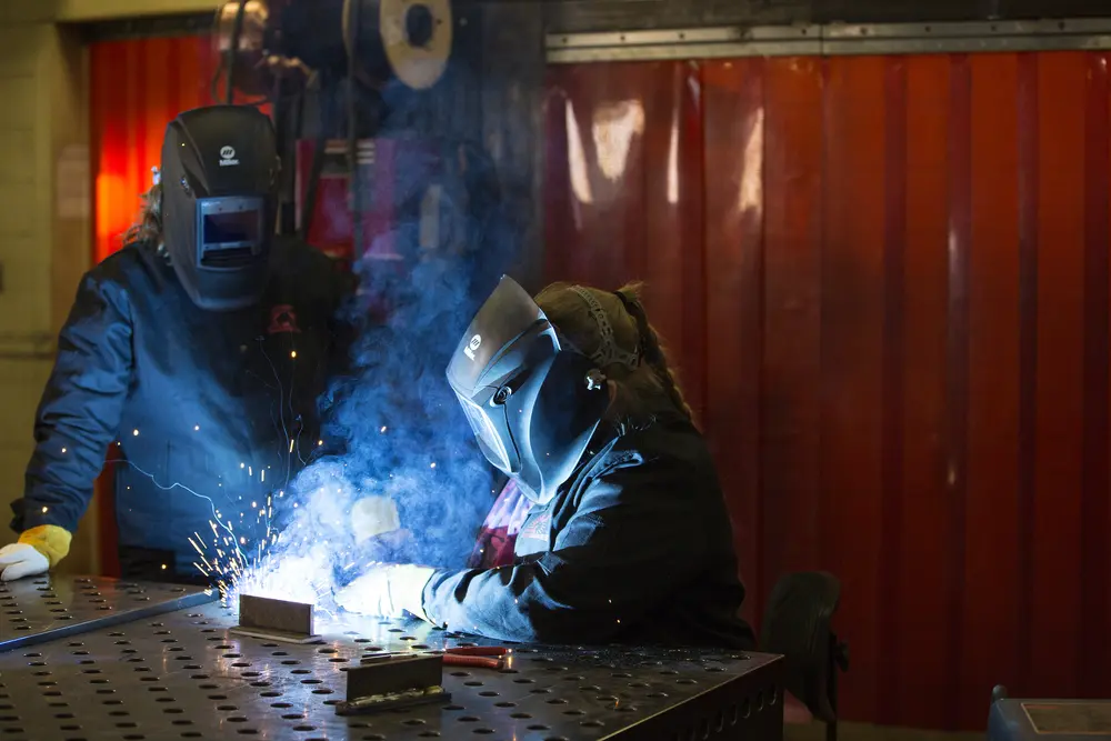 Two female welding students in protective gear work with bright sparks in a workshop.