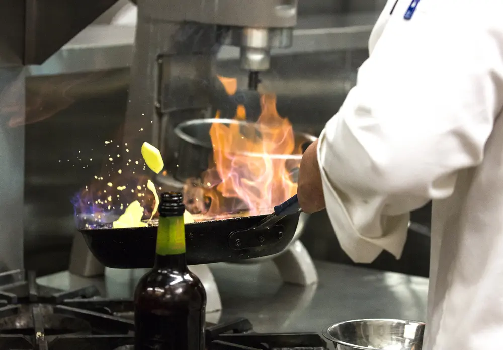 Culinary student flambéing a dish in a pan with flames on a stove.
