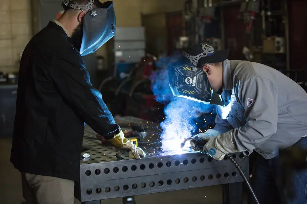 Welding student and instructor welding metal on a workshop table, sparks and blue light visible.