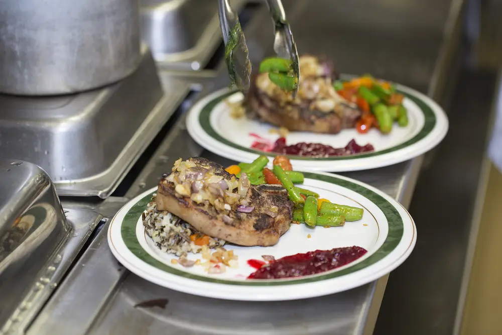 Pork chops with vegetables and sauce on white plates in a kitchen setting.