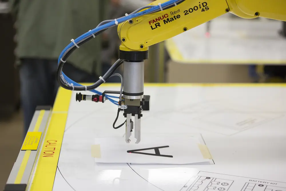 Yellow robotic arm writing a letter on paper with a pen attachment.