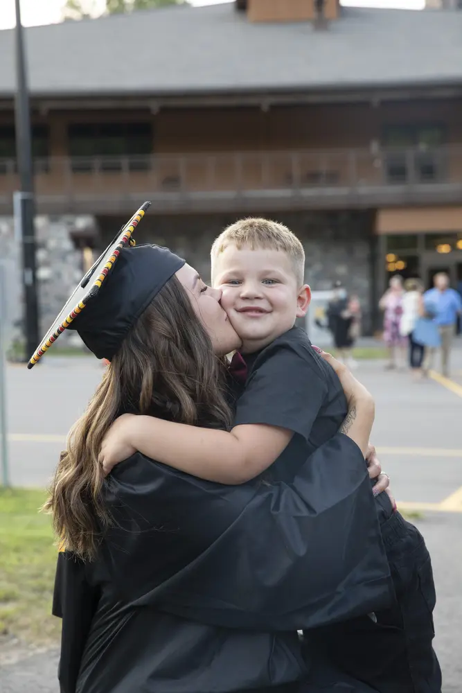 Mom graduate in cap and gown kisses smiling son on cheek outdoors.