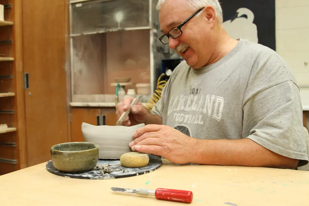 Older male student in a Nicolet College pottery class, smiling while sculpting a bowl.