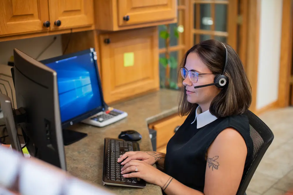 Female student with a headset working at a computer in an office setting.