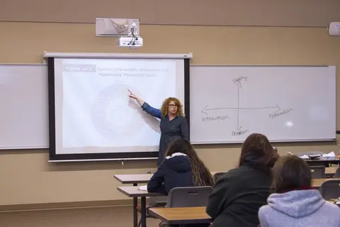 Female instructor pointing at a projector screen in a classroom with students.
