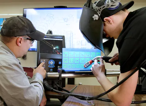Two people using welding simulators with a digital screen display.