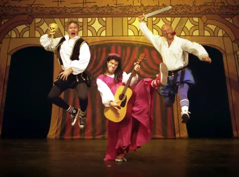 A group of actors on stage in Shakespearean costumes. Two are jumping. 
