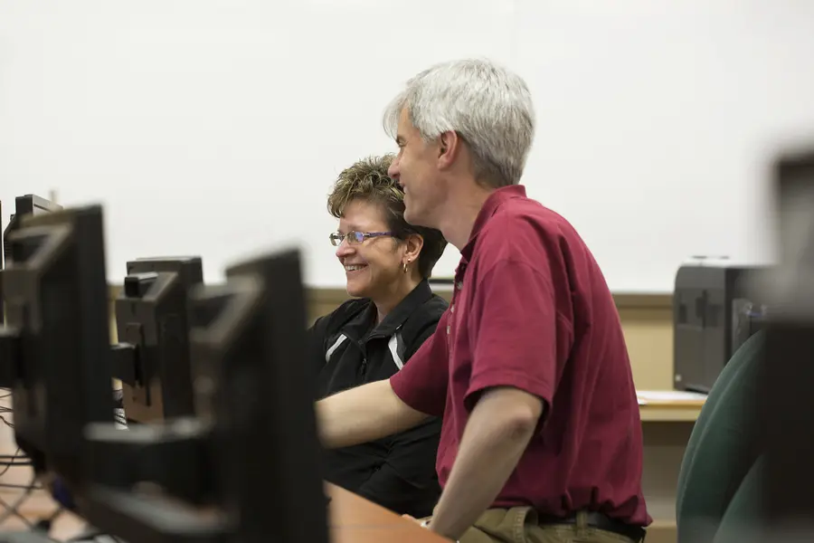 Accounting student with instructor, smiling and looking at computer screens in an office setting.
